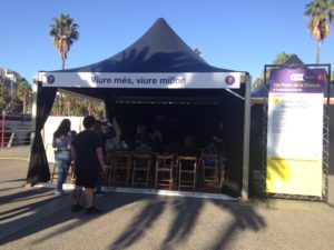 Image of a seating area at the event with Live more, live better tent on a sign above a tent around some wooden chairs people are sitting on watching something on a stage.